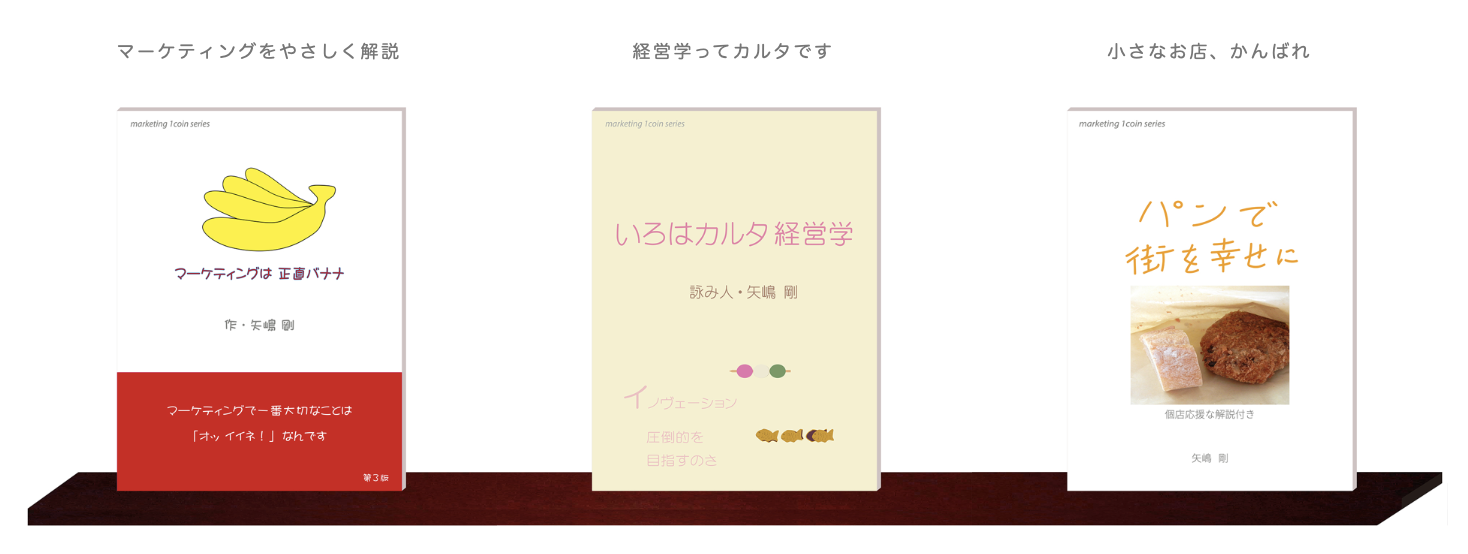 白い壁に取り付けたウォールシェルフの上に、こちらに表紙を向けて３つの本が並んでいます。『マーケティングは正直バナナ』『いろはカルタ経営学』『パンで街を幸せに：個店応援な解説付き』の表紙です。これらは矢嶋ストーリーから出ているマーケティング・１コイン・シリーズの第1巻、第2巻、第3巻です。著者は３冊とも矢嶋剛 Yajima, Takeshiです。
